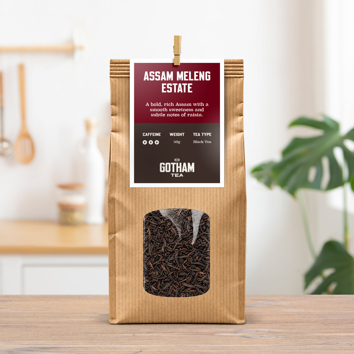 A 300g bag of Assam Meleng Estate black tea by Gotham Tea, in a brown kraft paper bag with a clear window showing the dark tea leaves inside. The bag is standing on a wooden table in a bright, modern kitchen setting.