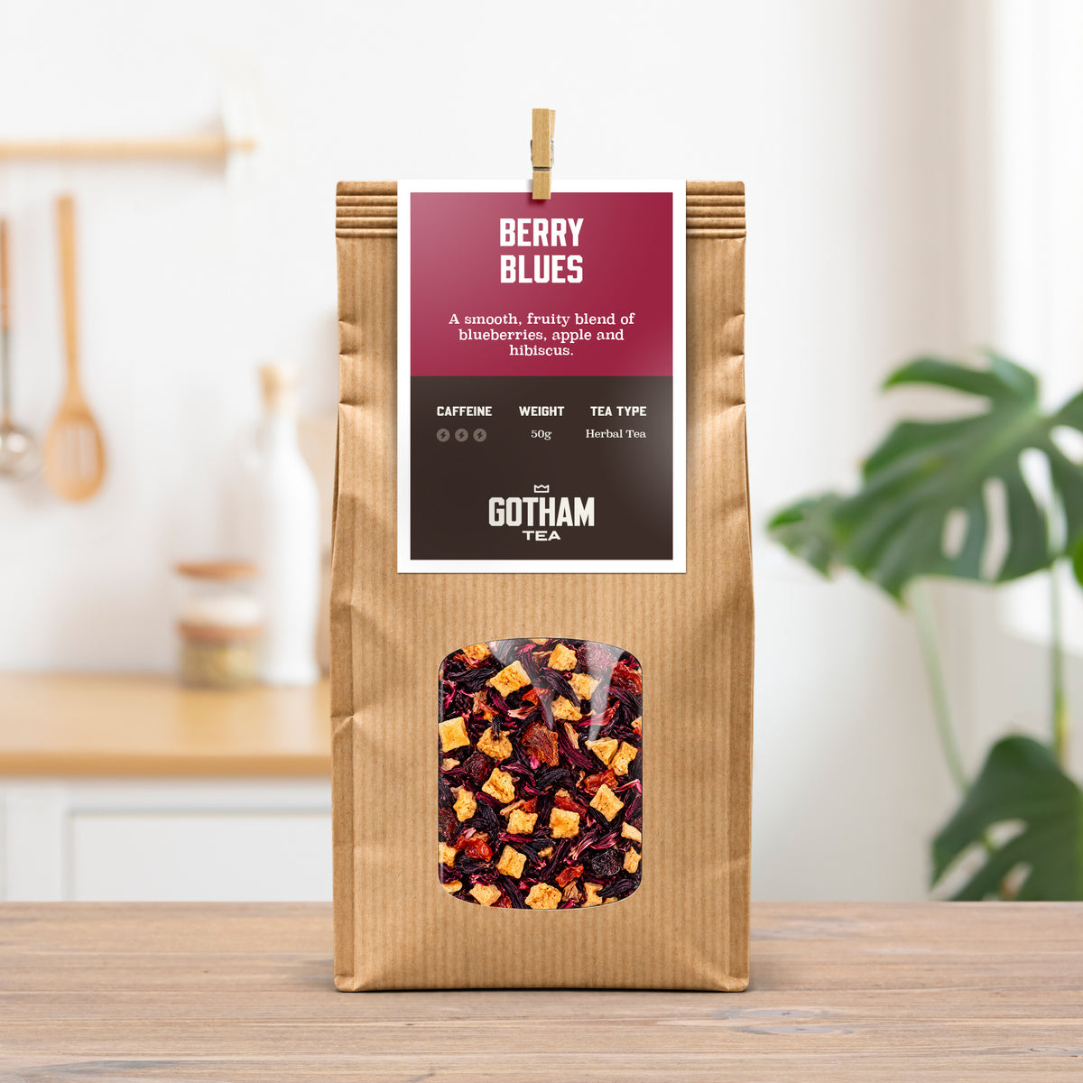 A kraft paper bag of Gotham Tea 'Berry Blues' herbal tea stands on a wooden table in a bright kitchen setting with a blurred plant in the background.