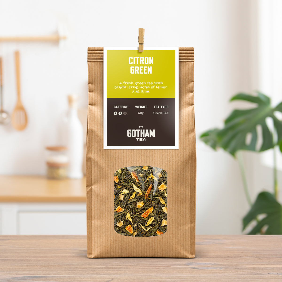 A bag of Gotham Tea Citron Green tea stands on a wooden table in a bright kitchen setting. The packaging is a brown kraft paper bag with a bright yellow-green and black label, and a clear window showing the loose-leaf tea inside.