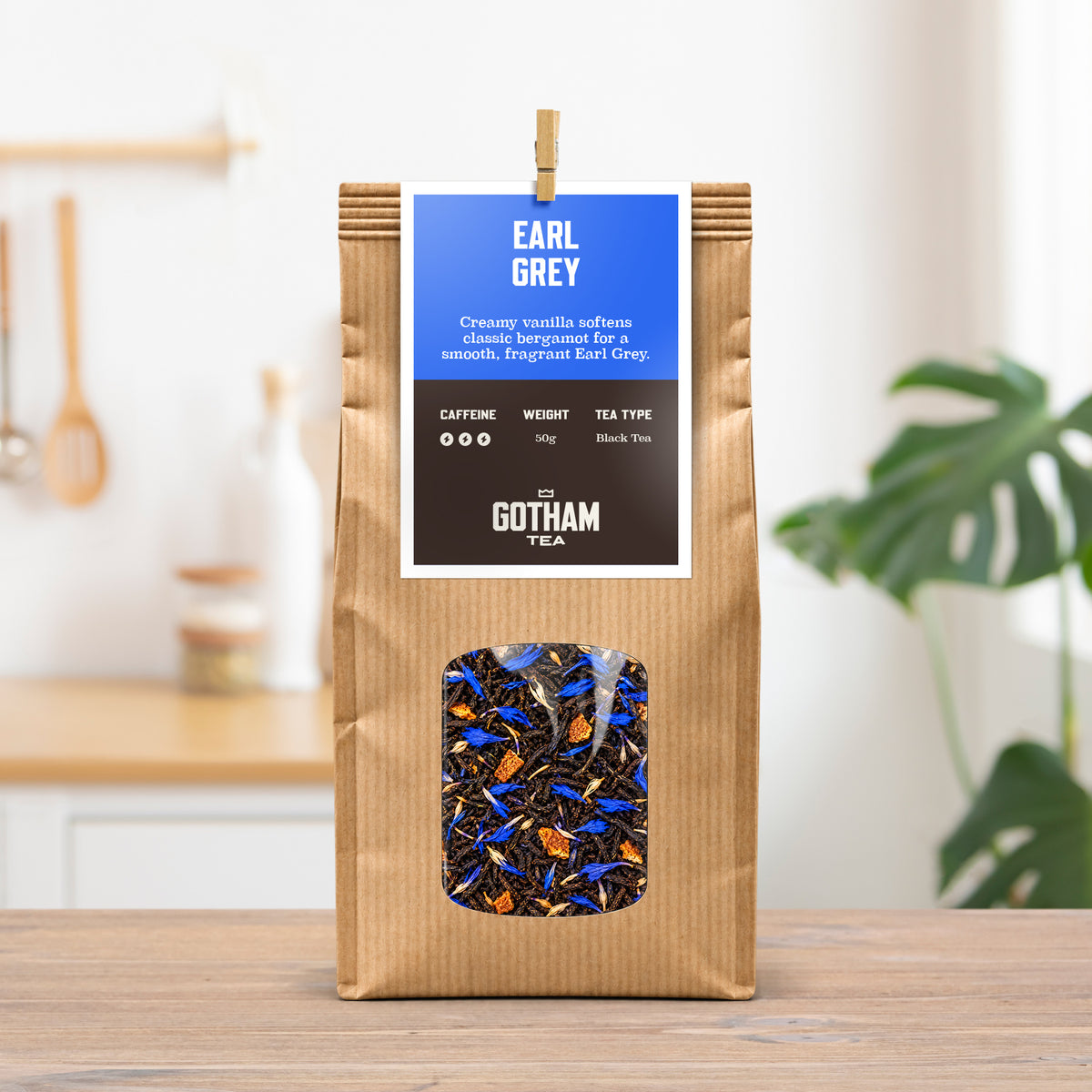 A kraft paper bag of Gotham Tea Earl Grey black tea stands on a wooden table in a bright kitchen setting. The packaging features a blue and brown label with a clear window showing the loose-leaf tea inside.
