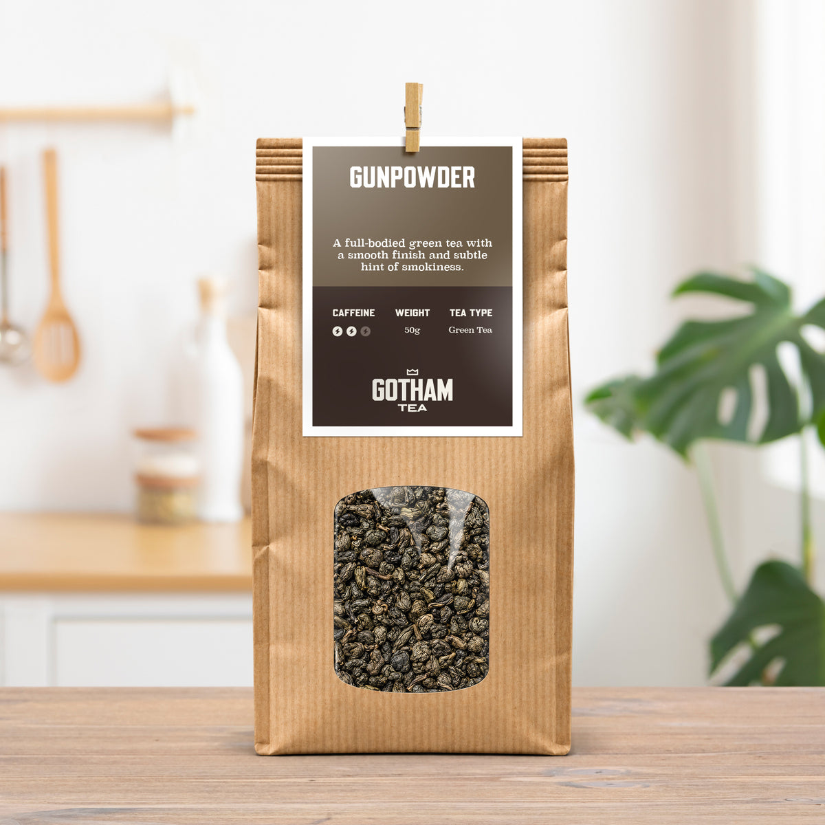 A bag of Gunpowder green tea from Tea by Gotham Tea, packaged in a brown kraft paper bag with a clear window showing the loose tea inside. The bag is set on a wooden table with a blurred kitchen background and a green plant.