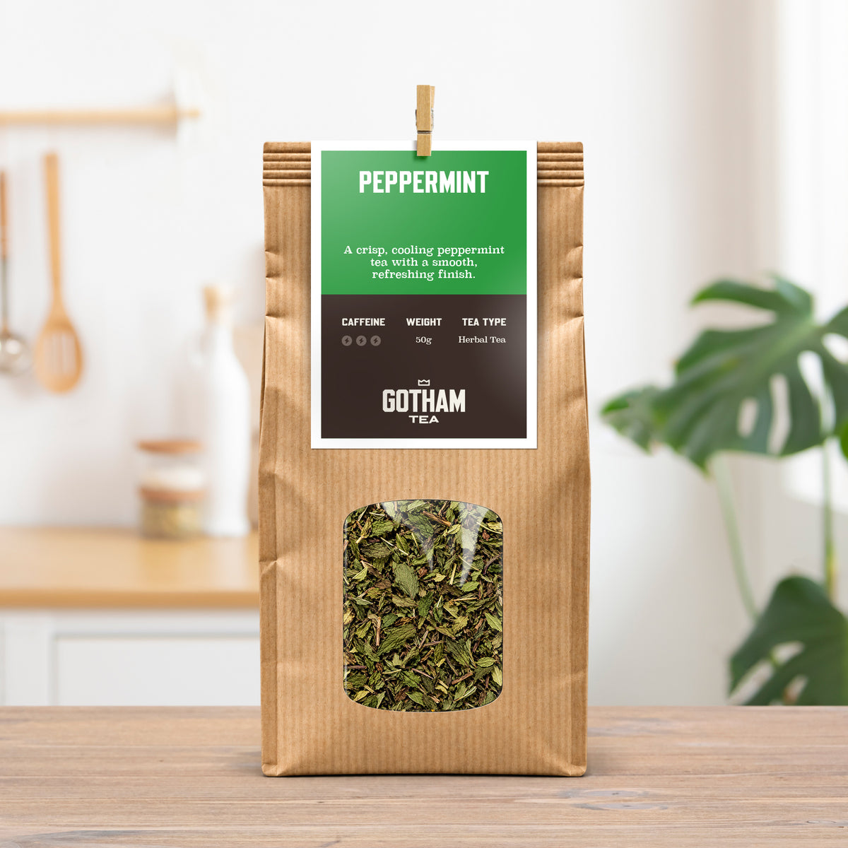 A kraft paper bag of Gotham Tea Peppermint herbal tea stands on a wooden table in a bright kitchen setting. The bag features a green and dark brown label with a clear window showing the loose green tea inside.